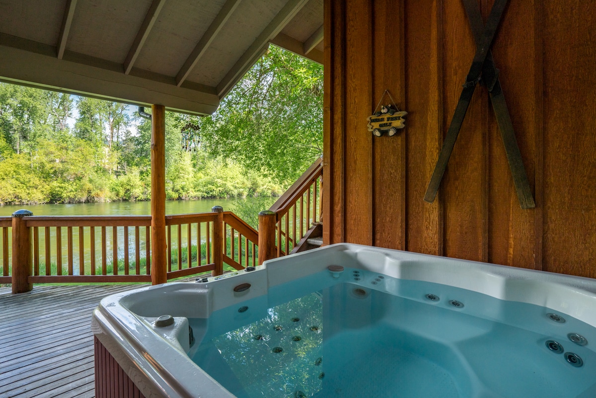 A private hot tub is situated on a wooden deck, offering serene views of the lush greenery along the Wenatchee River. Sturdy wood posts and a sloping roof provide shelter, creating a relaxing ambiance for guests to unwind.