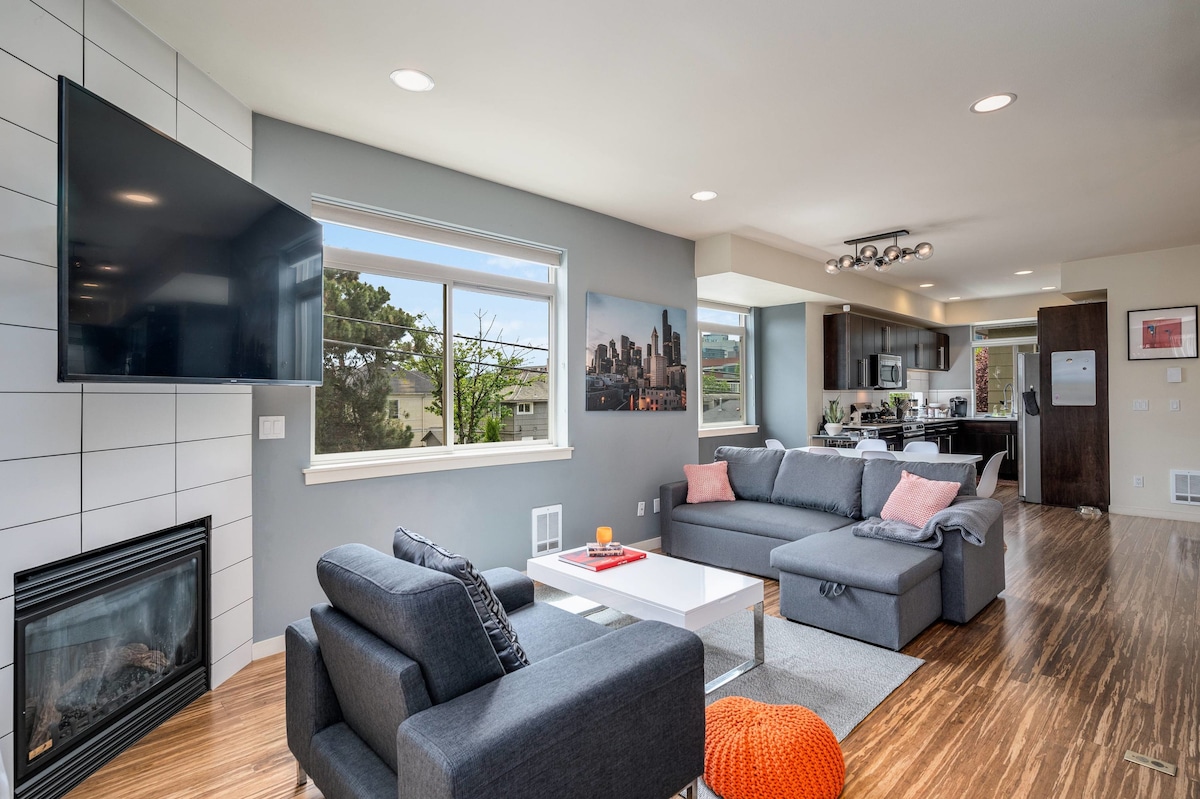 A modern living room is presented, featuring a flat-screen TV mounted above a white fireplace. Comfortable gray sectional sofas are arranged around a coffee table, which holds a red decorative tray. Natural light enters through large windows, offering views of greenery.