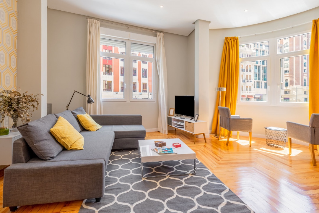A spacious living area is highlighted by large windows that provide abundant natural light. A gray sofa with yellow cushions complements a decorative area rug positioned under a coffee table. A television rests on a console, with two comfortable chairs placed beside the window.