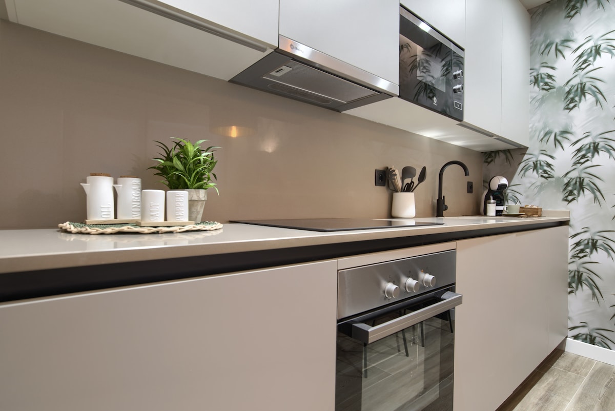 A modern kitchen area is highlighted, featuring sleek cabinetry and a built-in oven. A countertop is adorned with potted greenery and various kitchen accessories. The backdrop showcases a wallpaper with a botanical design, adding a touch of nature to the space.