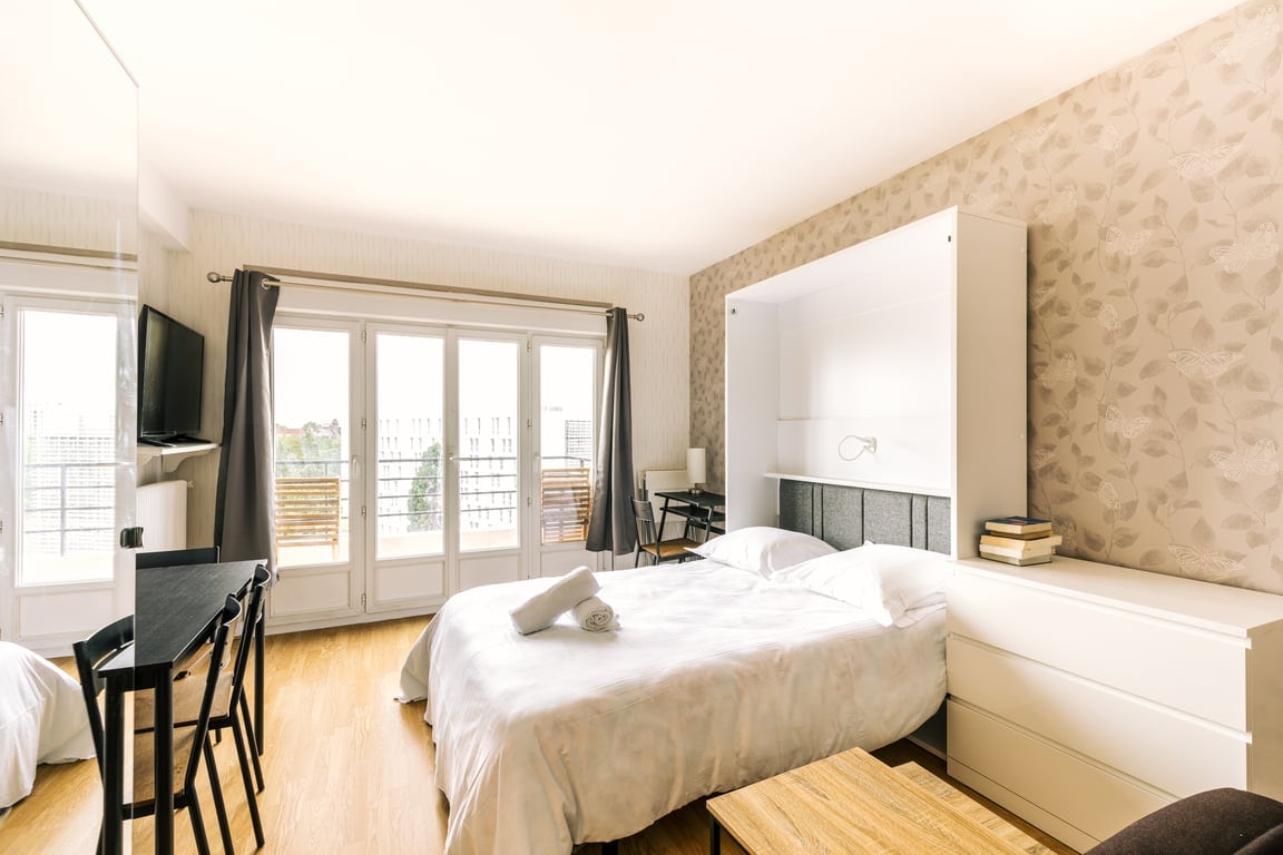 The spacious bedroom features a comfortable bed with fresh linens, complemented by a pair of modern chairs and a small table. Large windows offer ample natural light, and a mounted TV is positioned on the wall. A clean design is evident with soft color tones and decorative elements.