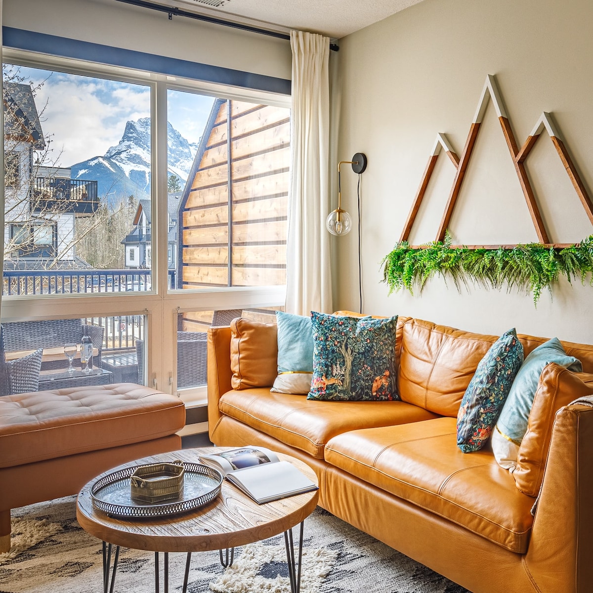 A cozy living area features a leather sofa adorned with colorful pillows. A circular table sits in front, accompanied by a book. Natural light floods in through large windows, showcasing mountain views beyond. Decorative elements include a geometric wall feature and greenery.