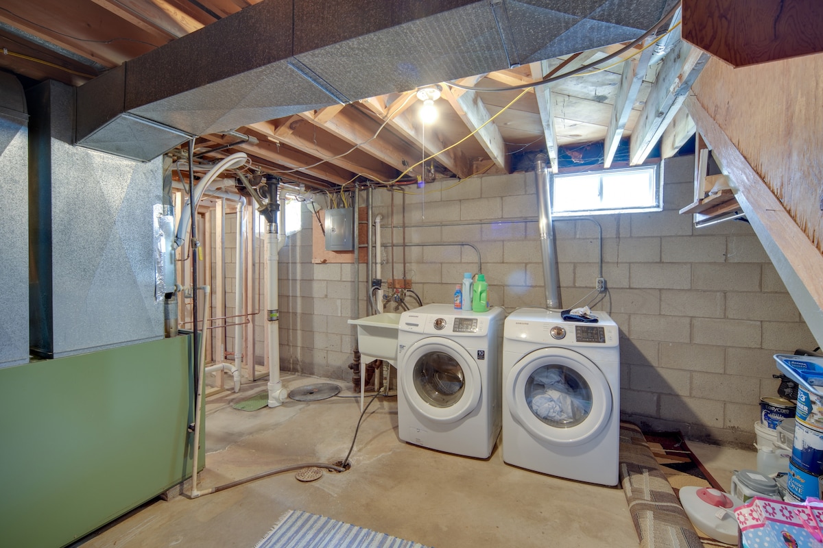 The laundry area features a pair of front-loading washing machines positioned beside a utility sink. Exposed wooden beams and ductwork are visible above, with light entering through a small window. The flooring is concrete, and various cleaning supplies are arranged nearby.