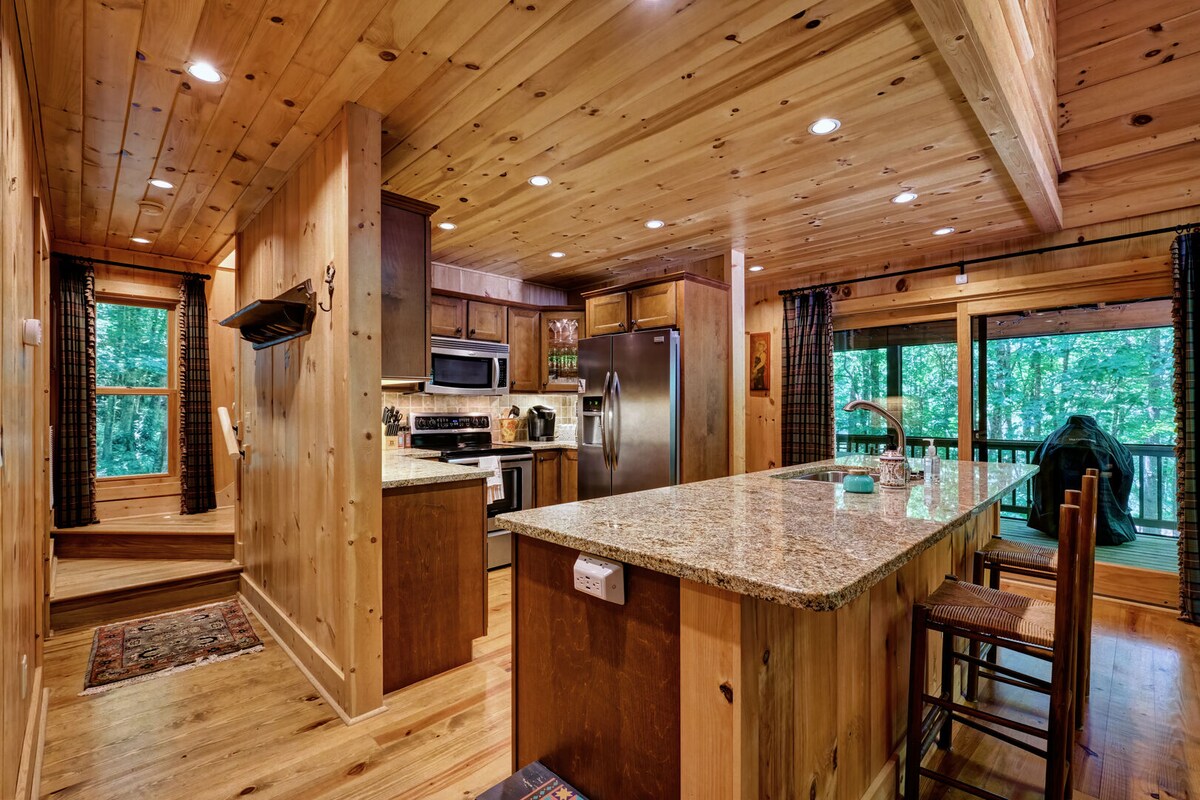 A wood-paneled kitchen area features modern appliances, including a stainless steel refrigerator and microwave. A spacious island with granite countertops is accompanied by bar seating. Natural light enters through large windows, revealing views of the wooded surroundings and access to an outdoor deck.