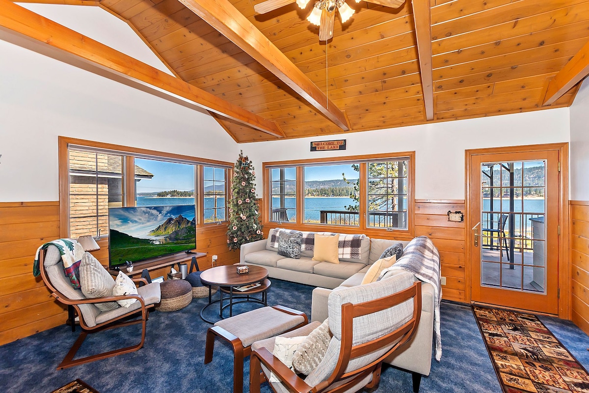 A cozy living area is highlighted by natural wood beams and a plush blue carpet. A large sectional couch faces a flatscreen TV, while a decorative tree adds a festive touch. Expansive windows reveal scenic views of Big Bear Lake and the surrounding landscape.