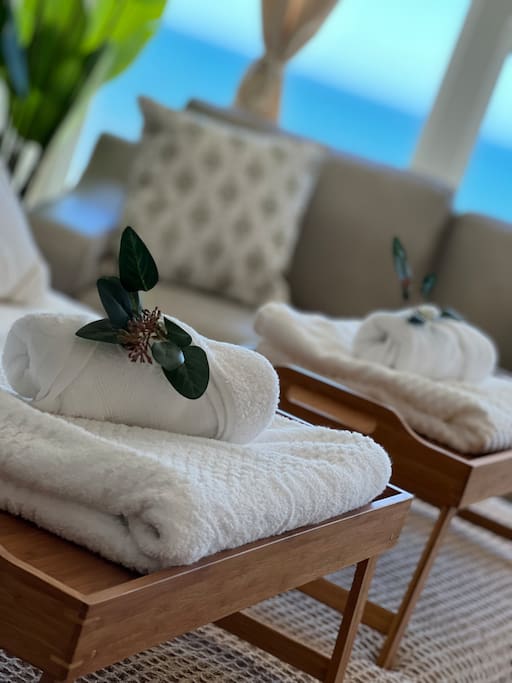 White towels are neatly rolled and placed on wooden side tables, accompanied by decorative greenery. A soft, couch is visible in the background, with a cushion adorned in a subtle pattern. Bright blue waters can be seen through large windows, enhancing the serene ambiance.