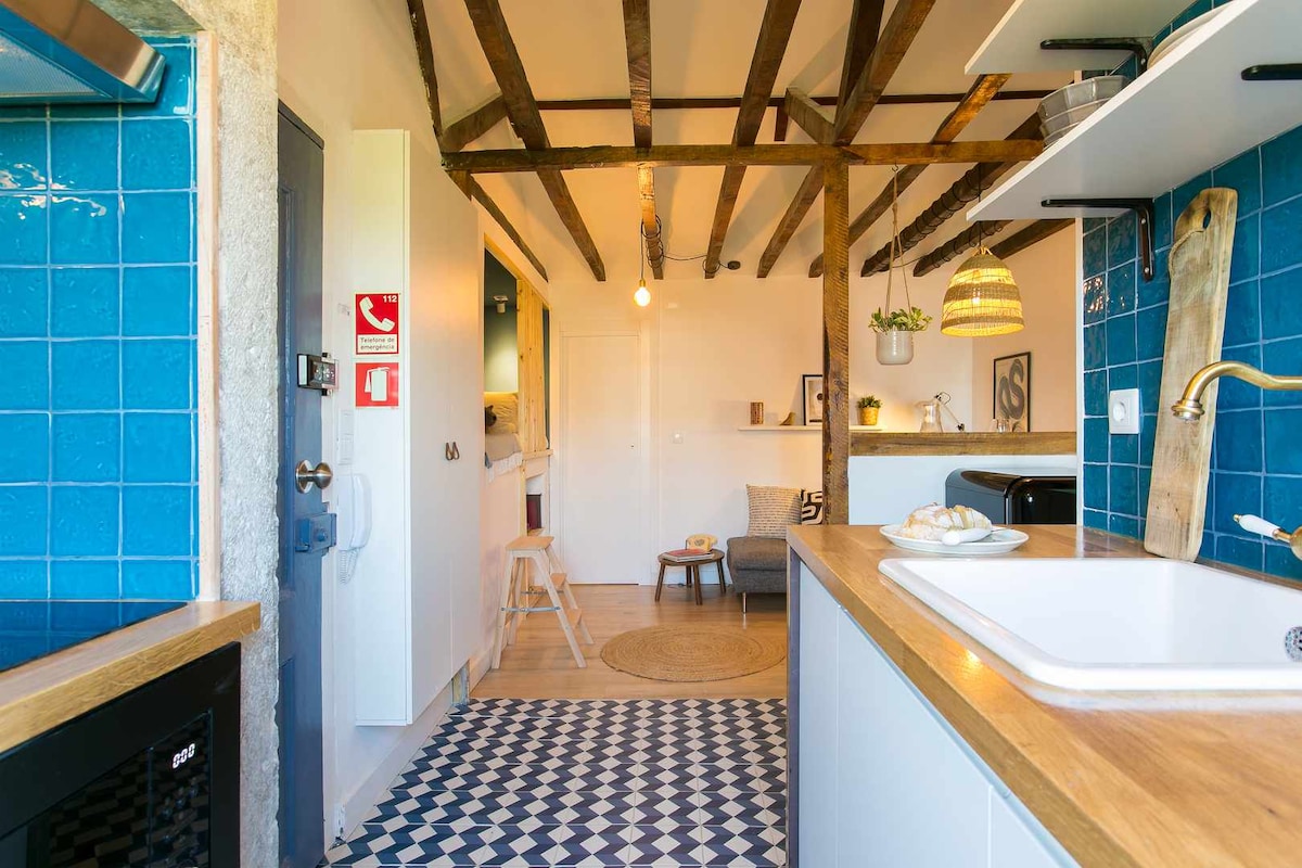 An open kitchen area features a white sink and wooden countertop, highlighted by textured blue tiles. Exposed wooden beams create visual interest above. A cozy seating area with a small gray sofa is visible in the background, with a circular rug placed on the floor.