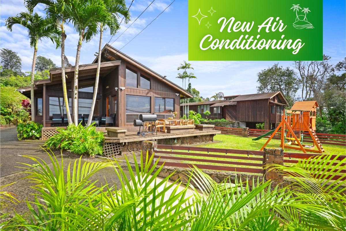 The exterior of the home features a wooden structure with large windows, surrounded by lush greenery. A spacious patio area includes seating, while a play structure can be seen in the background. Bright blue skies complement the tranquil setting.