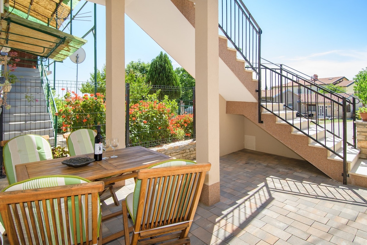 An outdoor terrace area features a wooden dining table with four chairs, accompanied by a bottle of wine. A staircase leads to an upper level, while blooming flowers and greenery enhance the ambiance, creating a pleasant space for relaxation.