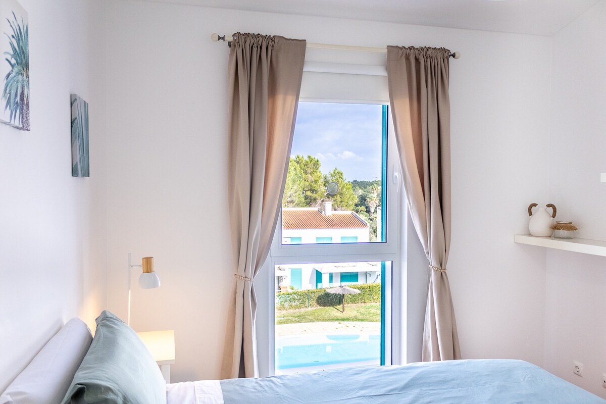 A bright and airy bedroom features a spacious bed next to a large window with soft curtains. A glimpse of the pool area and gardens is visible outside. A small shelf and a contemporary lamp add functional charm to the room's minimalist decor.