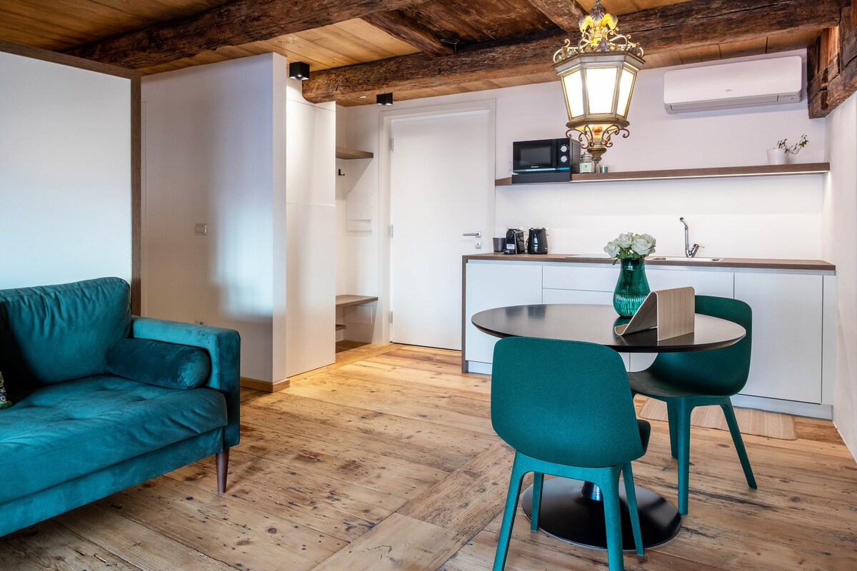 A cozy living space is presented with a green sofa and a round dining table surrounded by four matching chairs. A well-equipped kitchenette is visible, featuring modern appliances. Warm wood accents and flooring evoke a rustic charm, complemented by soft lighting from an overhead fixture.