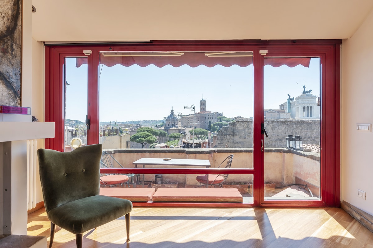 A large window provides expansive views over the historic sites of Rome, including the Forum and surrounding architecture. A cozy green chair sits near the window, complementing the inviting space. A small table and chairs are positioned on the terrace for outdoor dining.
