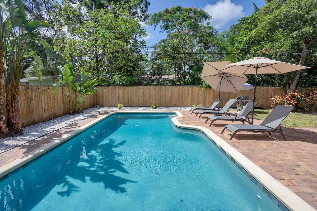 A fenced backyard features a heated swimming pool surrounded by four lounge chairs and two umbrellas. Lush greenery is present in the background, offering shade and a private feel. Sunlight reflects off the clear water, creating a serene outdoor space for relaxation.