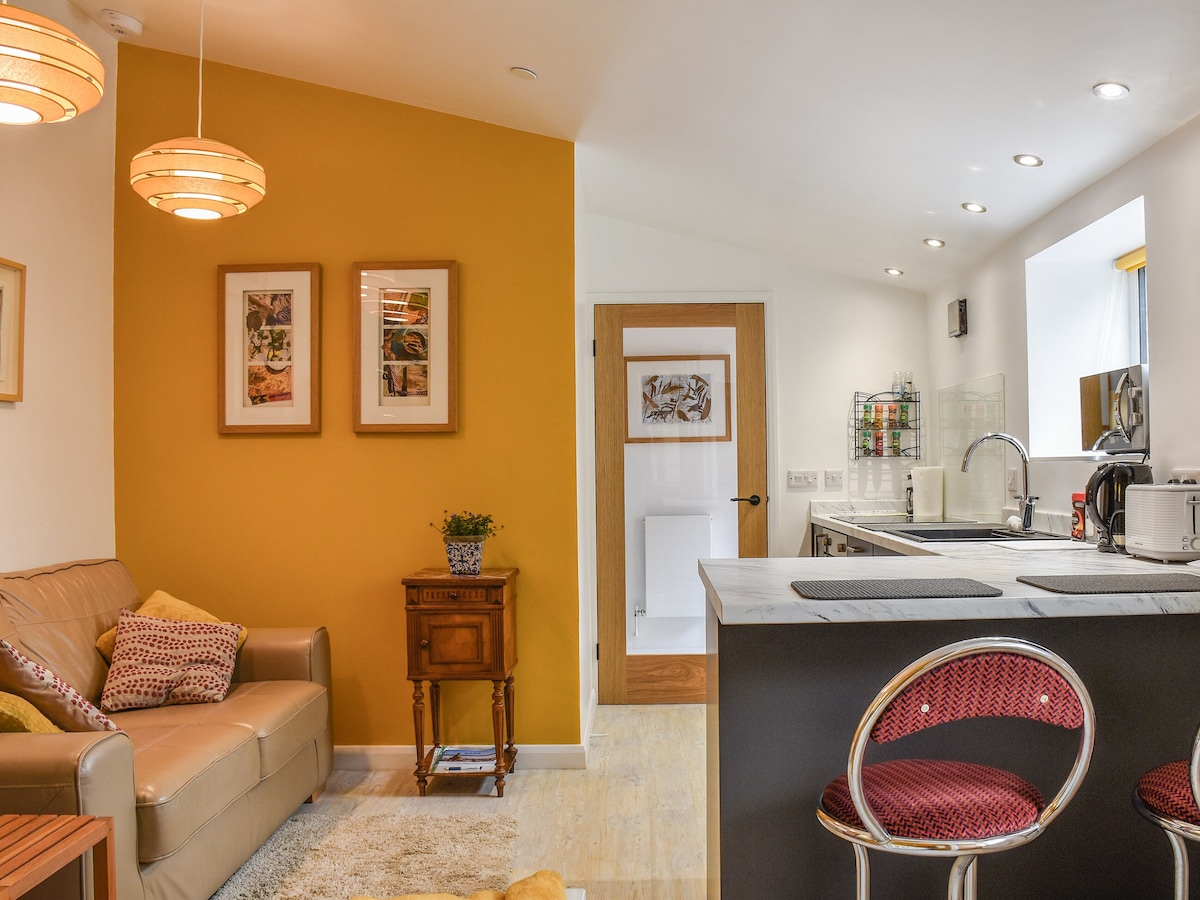 An open plan living space features a comfortable beige sofa adorned with patterned cushions, offering a cozy seating area. Two framed artworks hang on a golden wall, while a functional kitchen with modern appliances includes a breakfast bar highlighted by stylish red bar stools.