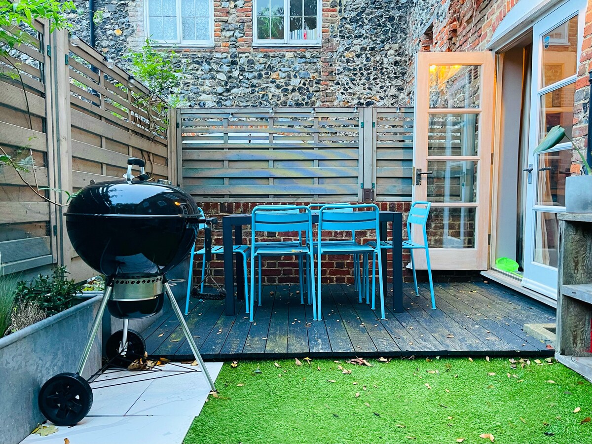 An outdoor patio is featured, showcasing a four-seater table with blue chairs and a nearby BBQ. Wooden decking surrounds the area, complemented by a grassy patch. The space offers a cozy setting for al fresco dining, bordered by rustic brick and wooden fencing.
