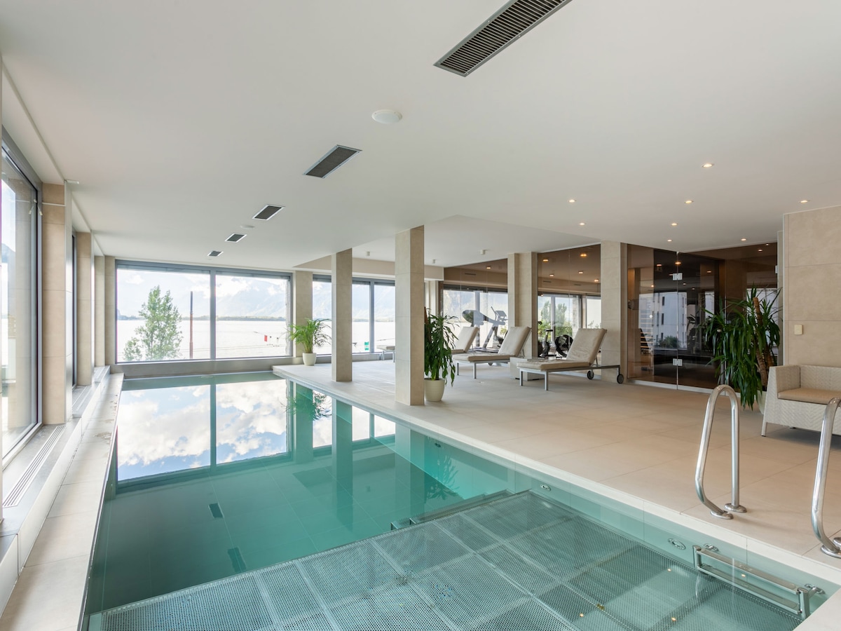 An indoor swimming pool area features a glass wall providing expansive views of the surrounding landscape. Sunlight reflects off the water's surface. Lounge chairs are positioned nearby, complemented by plants for a serene ambiance. The space is bright and open, highlighting a modern design.