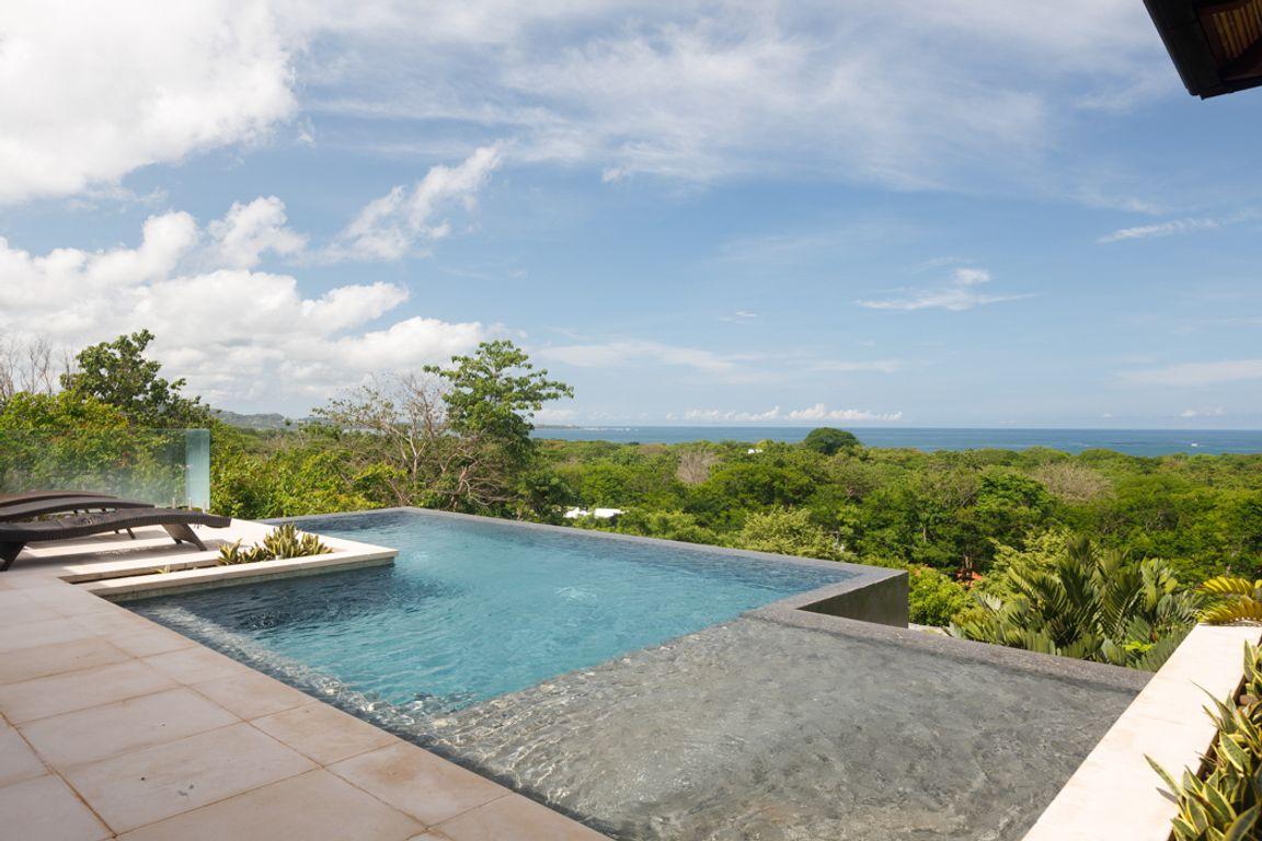 An infinity edge pool is seen overlooking a lush green landscape, with views of the Pacific Ocean in the distance. The pool area is surrounded by tropical vegetation and features a sun lounger positioned for relaxation under the clear blue sky.