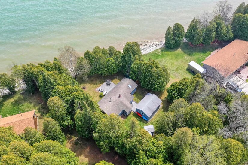 An aerial view captures the Hidden Shores Cottage nestled among lush greenery alongside Lake Michigan. The expansive yard features a mix of mature trees and open space leading to the water, creating a secluded atmosphere perfect for relaxation.