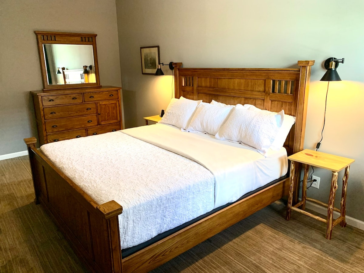 A king size bed is centered against a muted wall, framed by two bedside tables with lamps. A wooden dresser with a mirror is positioned nearby, adding to the room's warm and natural aesthetic. Soft light illuminates the space, enhancing comfort.