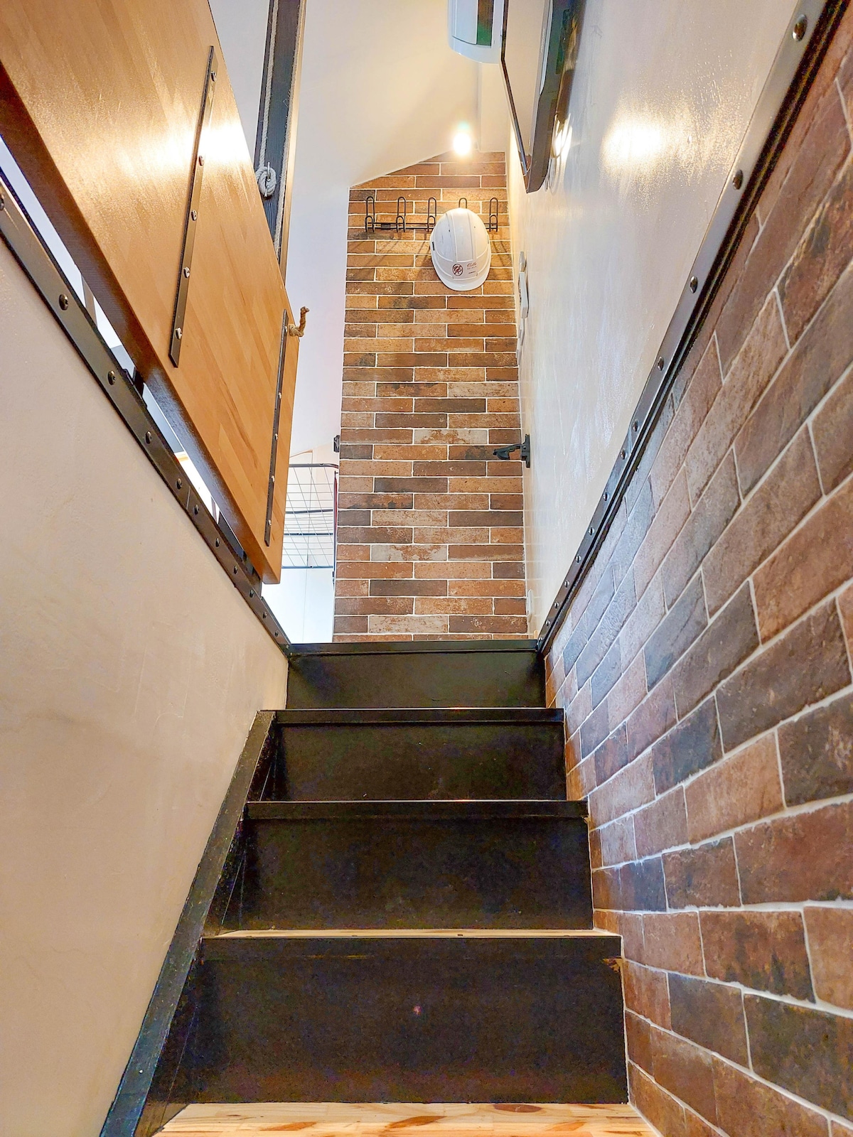 A staircase with black steps leads upward, flanked by exposed brick walls. Metal railings provide support along the side. Soft lighting illuminates the area, enhancing the modern design. A small window is visible at the top, adding a touch of natural light to the space.