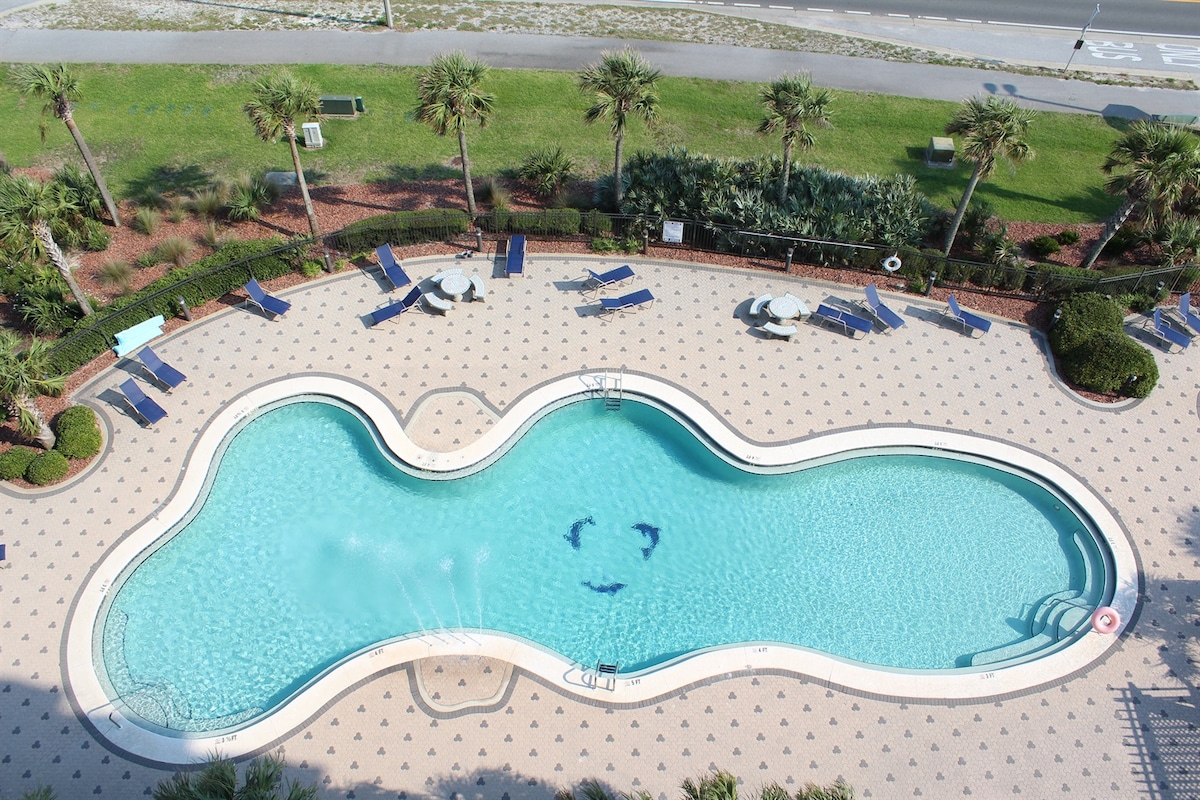 A spacious outdoor pool is depicted, surrounded by lounge chairs and greenery. The pool features a unique, organic shape and is bordered by a textured deck. Several tables with umbrellas are positioned nearby, providing shaded seating areas for relaxation.