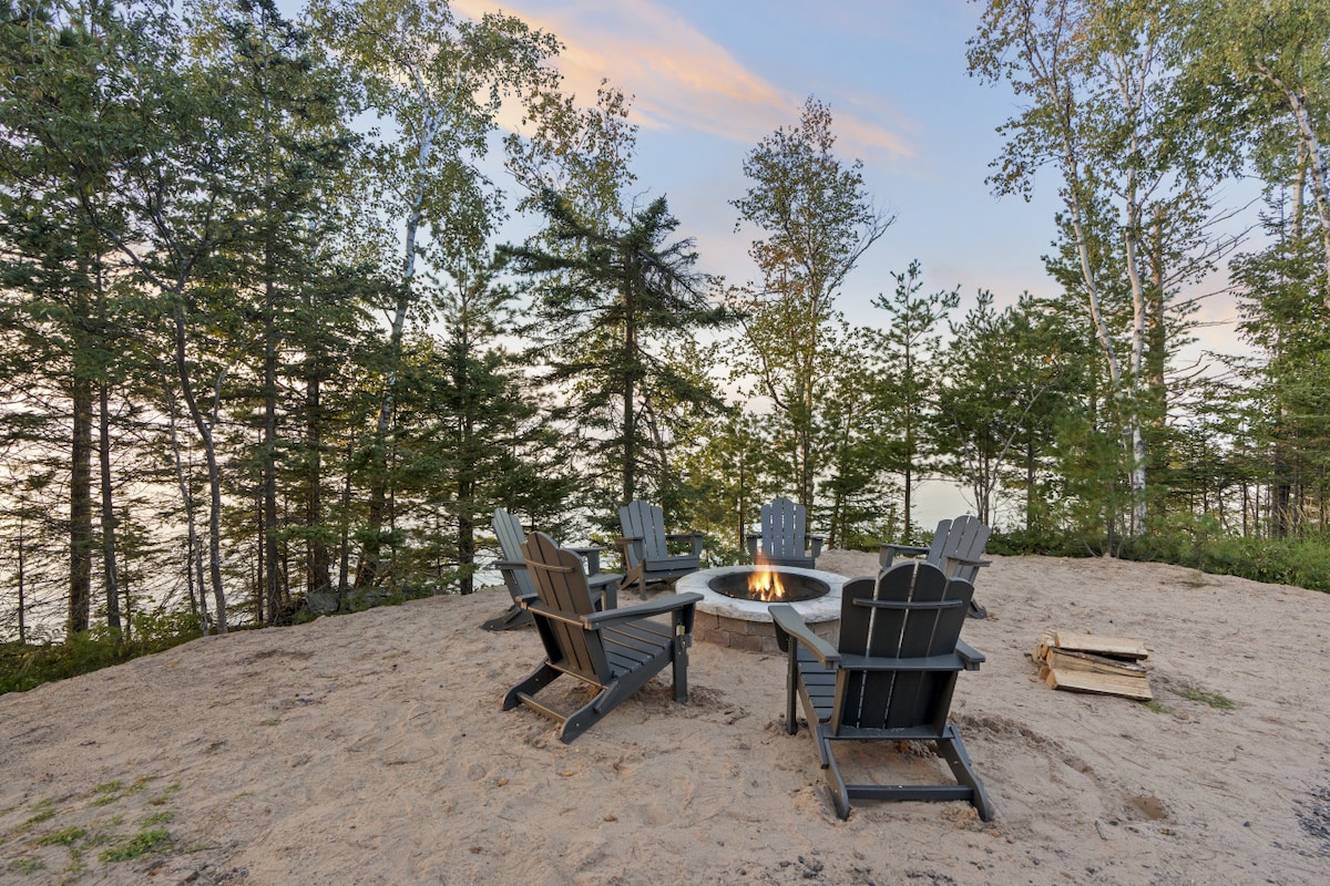 A sandy firepit area is surrounded by five black Adirondack chairs, providing a space for gatherings. The backdrop features trees that frame the scenic view of the shoreline, with the calm waters of Lake Superior visible in the distance.