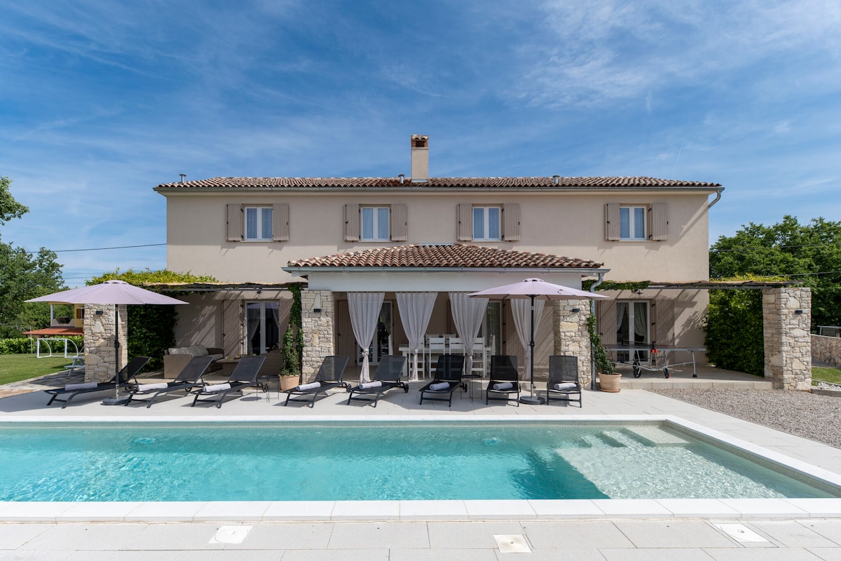 The spacious villa is depicted with a charming façade, featuring light-colored walls and a tiled roof. Sun loungers are arranged beside the clear outdoor pool, and shaded areas are created by large umbrellas. Lush greenery surrounds the property, inviting relaxation.
