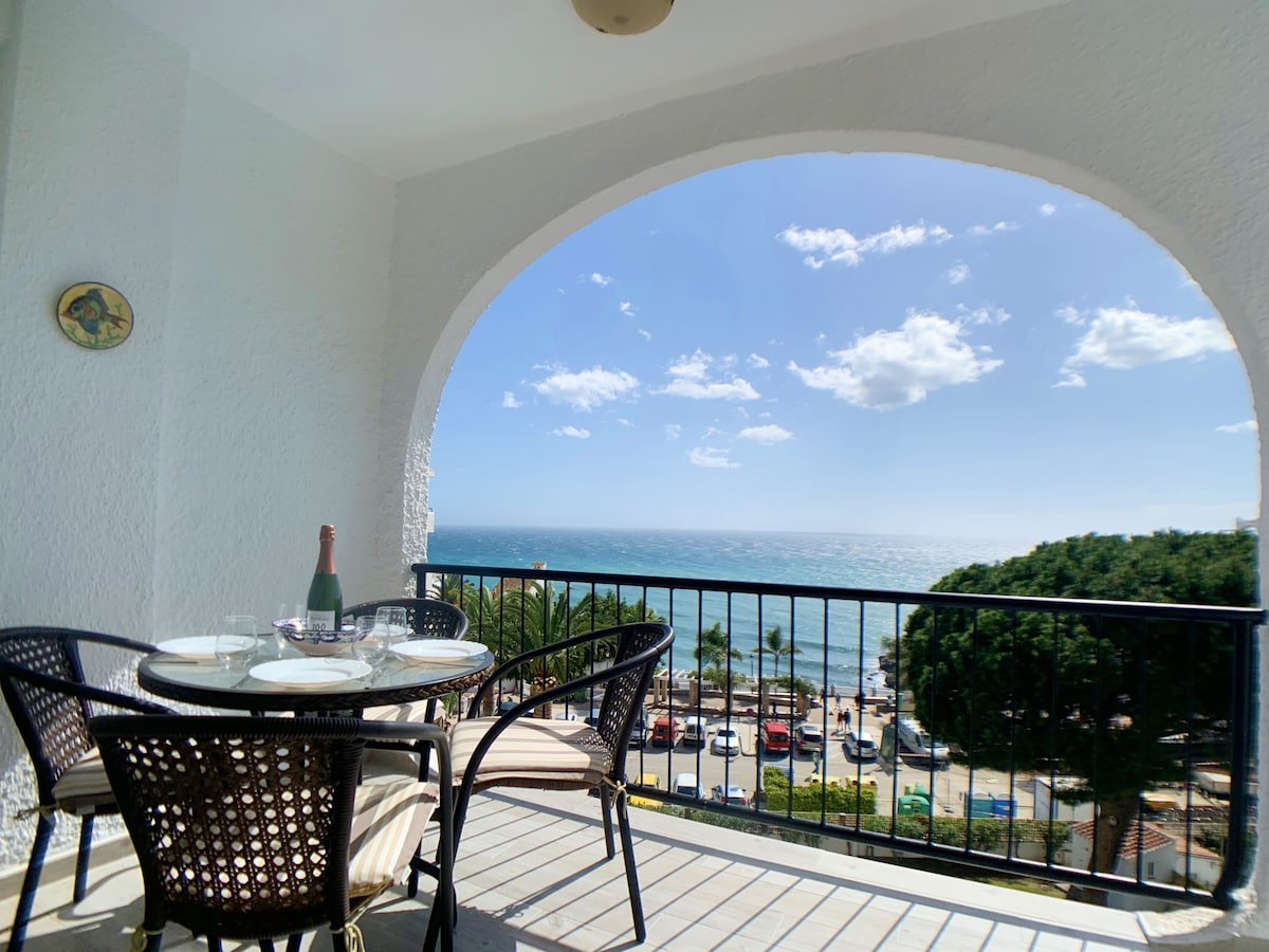 An inviting terrace features a round table set for four, accompanied by glasses and a bottle. The sea view is framed by an arched opening, highlighting the horizon and a partially cloudy sky. Lush greenery and parked cars are visible along the beachfront.