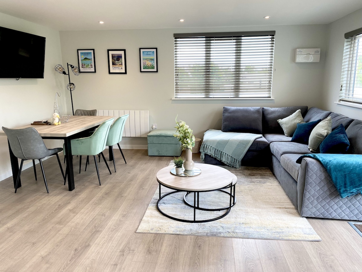 An open-plan living area features a comfortable gray sofa, a wooden dining table, and stylish chairs. Framed pictures adorn the walls, and large windows provide natural light. A round coffee table is centered on a soft area rug, enhancing the inviting space.