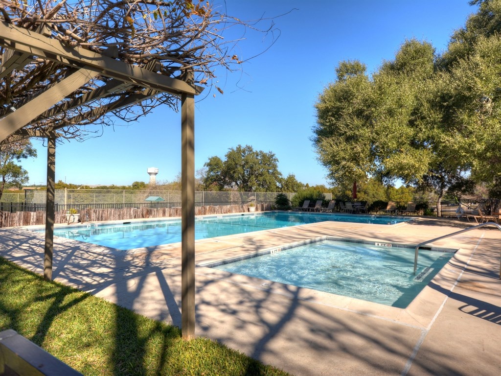 A clear view of a swimming pool area featuring a spacious main pool and a smaller hot tub. Surrounding lounge chairs are arranged along a concrete deck, complemented by vibrant green grass and shaded by surrounding trees.