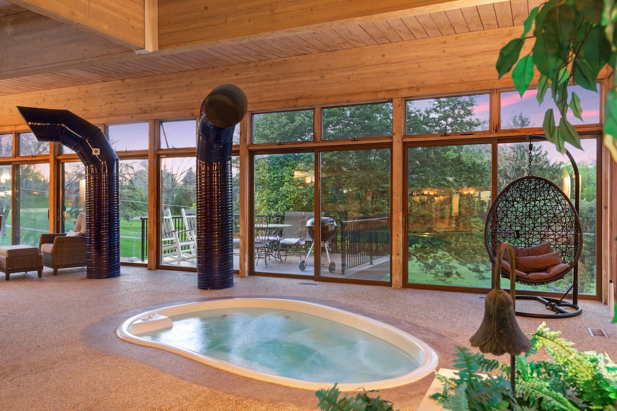 An indoor hot tub is showcased in a spacious area with floor-to-ceiling windows providing views of the lush outdoors. Natural light fills the space, enhancing the tranquil ambiance. A hanging chair and comfortable seating invite relaxation, creating a serene setting for unwinding.
