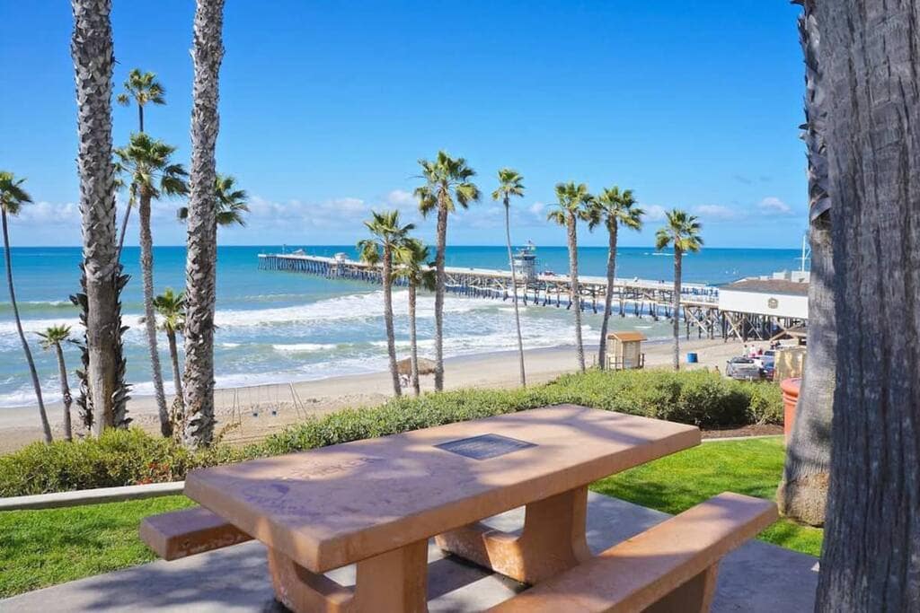A concrete picnic table is positioned in a grassy area, overlooking a sandy beach and ocean waves. Palm trees frame the view, which includes a pier extending into the water under a clear blue sky.