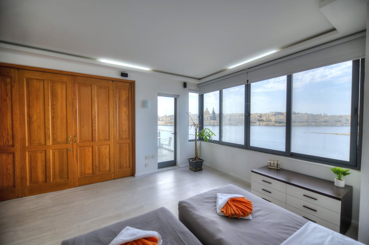 The spacious bedroom features large windows offering views of the sea and Valletta. Wooden closet doors add warmth to the decor, while a dresser is positioned adjacent to the beds. Two single beds are neatly arranged with orange towels placed on each, creating a simple yet inviting atmosphere.