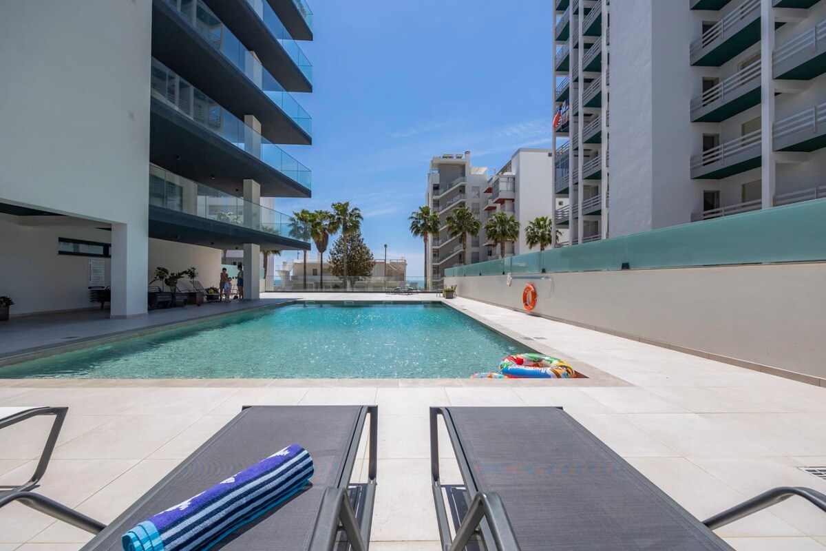 A communal pool is framed by modern buildings, with two sun loungers positioned by the water's edge. Brightly coloured towels are neatly placed on the loungers, and a float can be seen in the pool. Palm trees line the background, complementing the sunny atmosphere.
