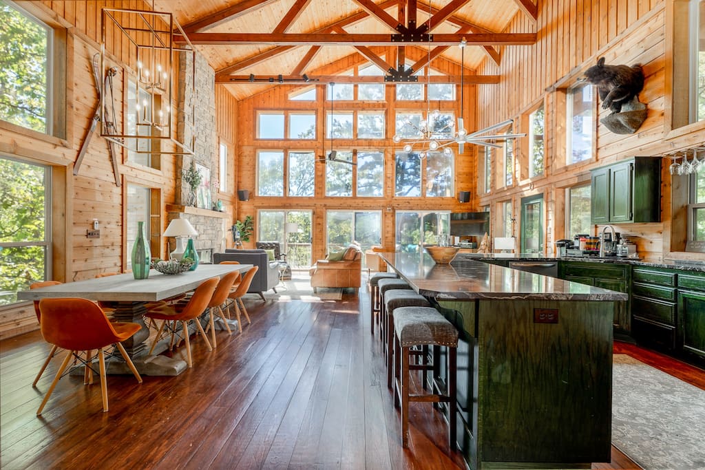 An open-concept living area features high vaulted ceilings with wooden beams and large windows allowing ample natural light. A dining table and spacious kitchen island are accompanied by modern seating and rustic decor, creating a welcoming environment for gatherings.