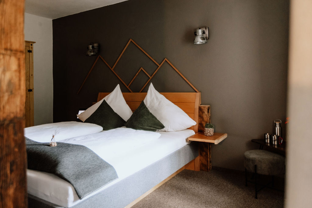 A spacious bedroom features a wooden bed with two large pillows adorned with earthy-toned covers. Soft green accent pillows are placed on the bed, complementing the light grey bedding. A textured wall art piece resembling mountains is mounted above the headboard, while warm lighting is provided by wall sconces.