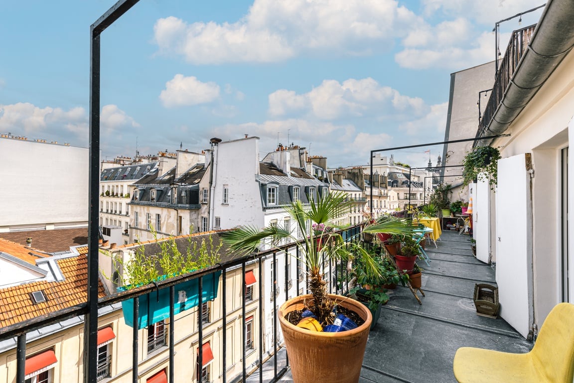 A balcony offering a view of the surrounding rooftops of Paris is highlighted by potted plants and colorful furniture. The spacious area features a railing, allowing for an open breeze, enhancing the outdoor experience.