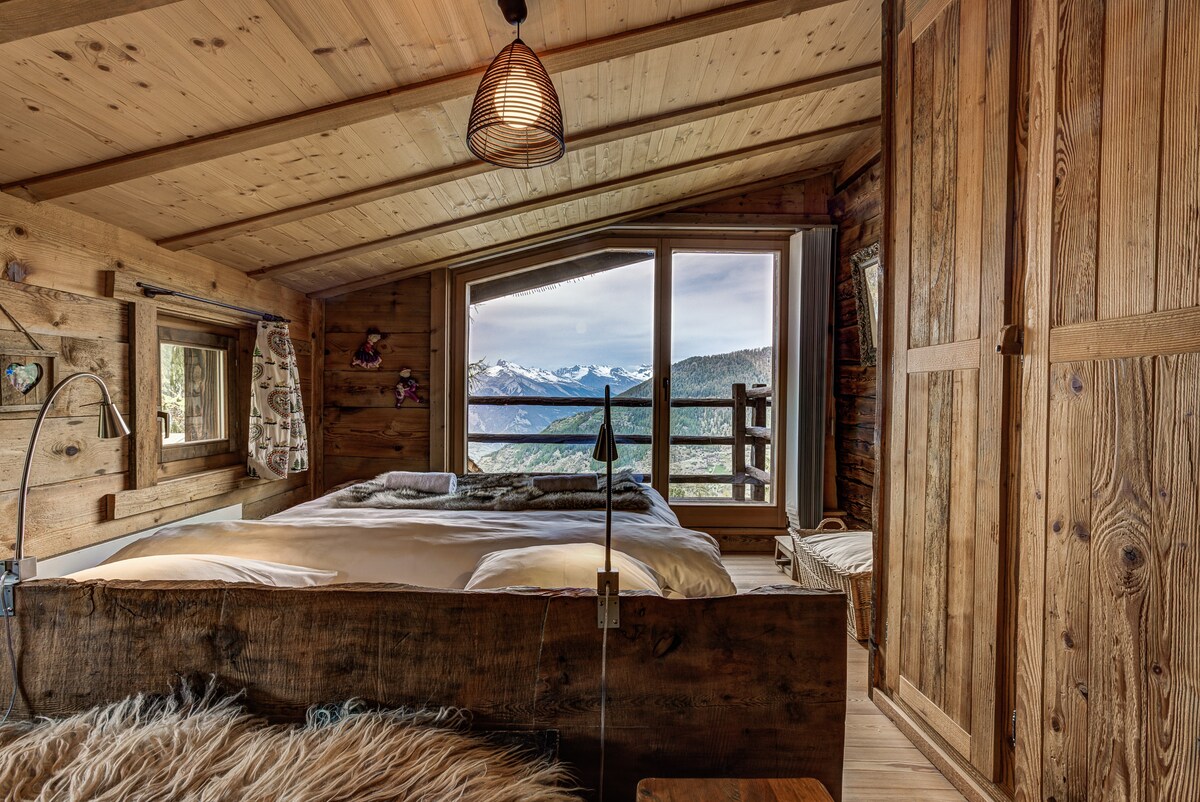 A cozy bedroom features a spacious bed surrounded by rustic wooden walls and ceiling. Large windows offer panoramic views of the mountains, allowing natural light to fill the room. A bedside lamp and woven light fixture add warmth to the serene atmosphere.