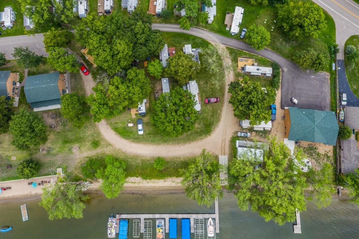An aerial view showcases a campground area bordered by greenery and a waterway. A winding road is visible, leading to various cabins and parking spots. Boating facilities and a dock extend into the water, indicating opportunities for outdoor activities.