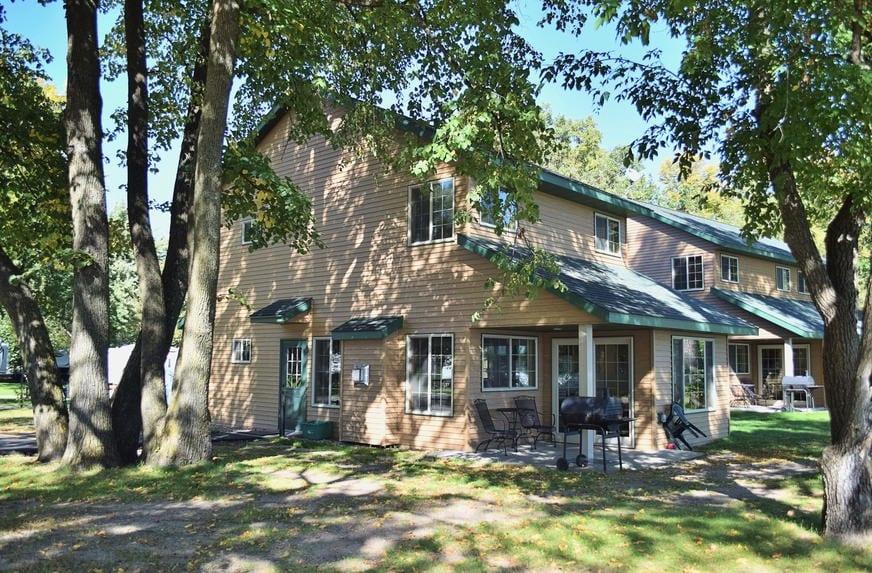 A charming two-story cabin is surrounded by trees, featuring a natural wooden exterior and large windows. A patio area is visible with seating and a grill, inviting outdoor gatherings. The setting is peaceful, highlighted by soft sunlight filtering through the leaves.