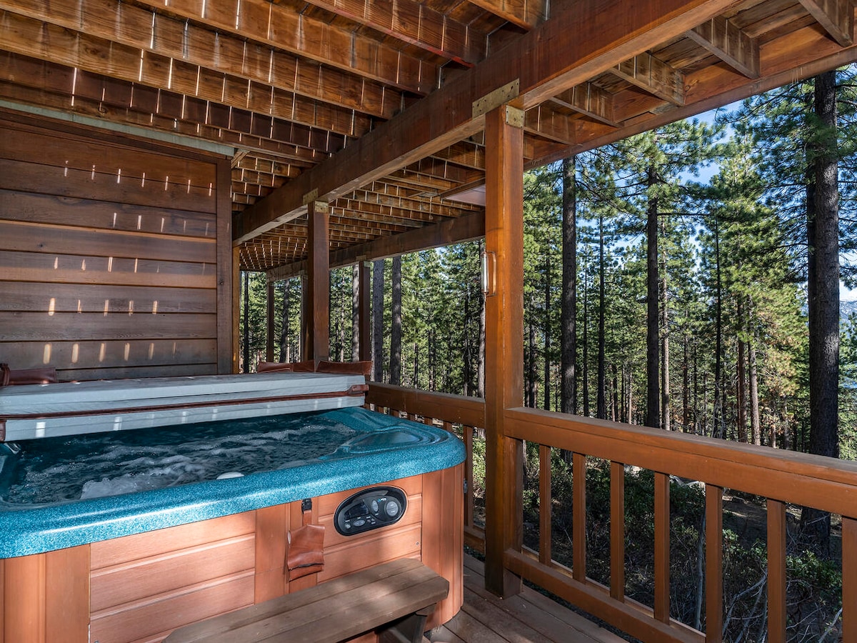 A hot tub is situated on a wooden deck, surrounded by wooden balustrades. The peaceful forest backdrop showcases tall green trees, creating a secluded atmosphere. Natural light filters through the space, enhancing the inviting setting for relaxation.