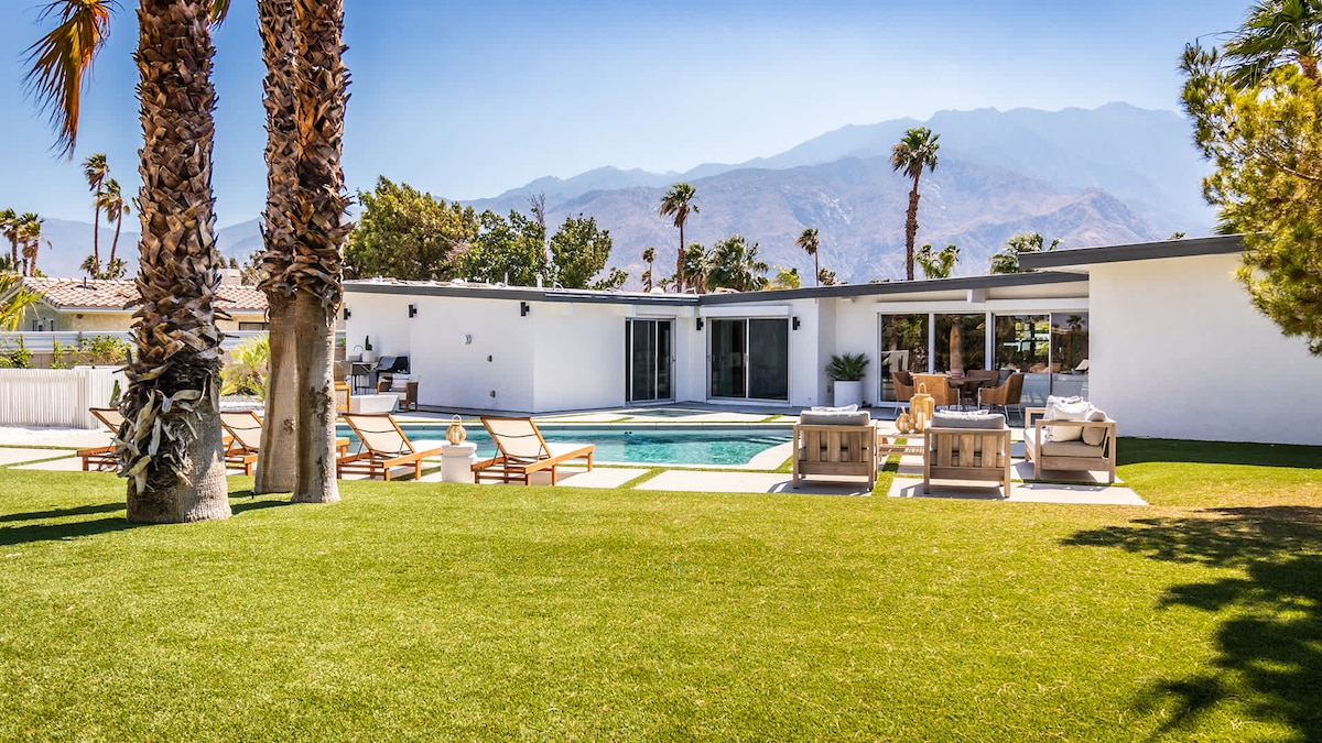 A spacious backyard is highlighted by a crystal-clear pool surrounded by lounge chairs. The villa's modern exterior features large glass doors, providing a seamless transition to the indoor spaces. The San Jacinto mountains rise in the background, enhancing the serene landscape.