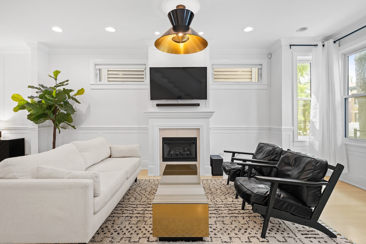A modern living room features a light-colored sectional sofa facing a sleek coffee table. Two black armchairs flank the table, and a wall-mounted television is positioned above a decorative fireplace. Large windows bring in natural light, complemented by minimalistic decor.