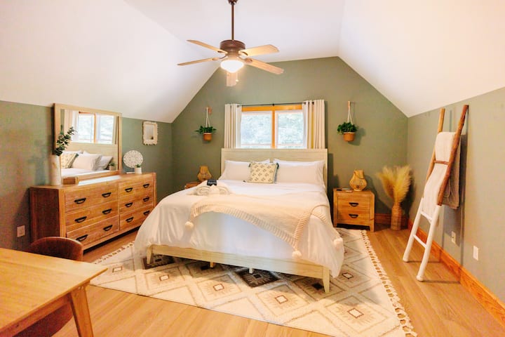 Ბოჰემური სტილის მთავარი საძინებელი დიდი ზომის საწოლით, ბუნებრივი მცენარეებით, სრული ზომის სარკითა და მაგიდით.