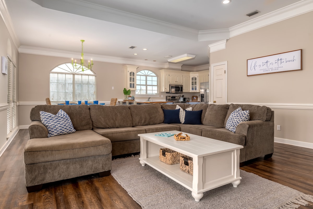 A spacious living area features a large sectional sofa adorned with decorative blue and white cushions. A white coffee table rests in the center, paired with woven storage baskets. The room is illuminated by natural light from multiple windows and has a view of the adjacent kitchen.