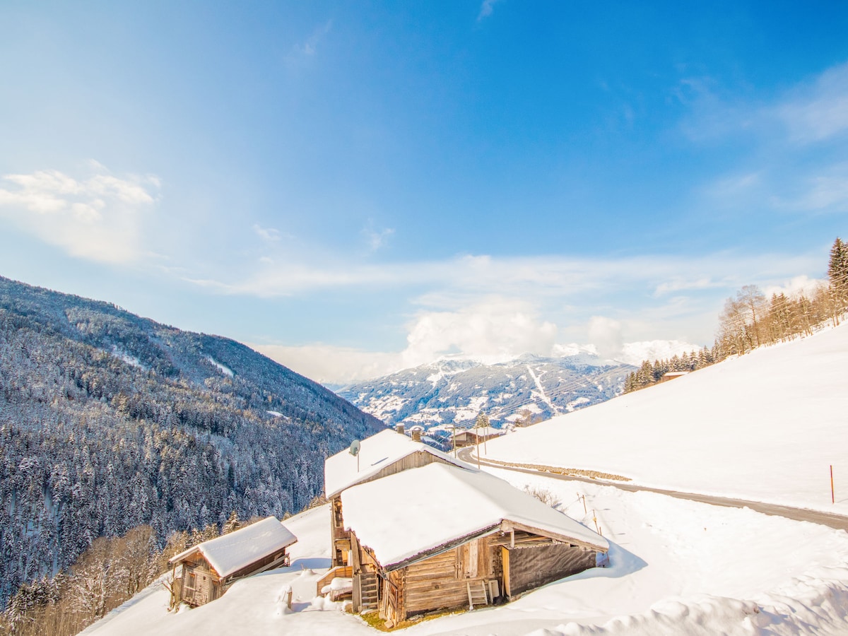 A serene winter landscape features traditional wooden structures blanketed in snow. Majestic mountains rise in the background, framed by clear blue skies. The pathways, lined with snow, invite leisurely strolls. The overall scene reflects the tranquility of a snowy countryside.