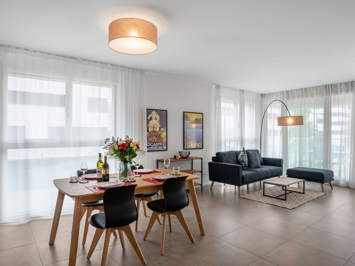 An open living/dining area is presented with a modern dining table set for four and a comfortable sofa. Large windows draped in sheer curtains allow natural light to illuminate the space, creating a bright and airy atmosphere. Decorative artwork is displayed on the walls.