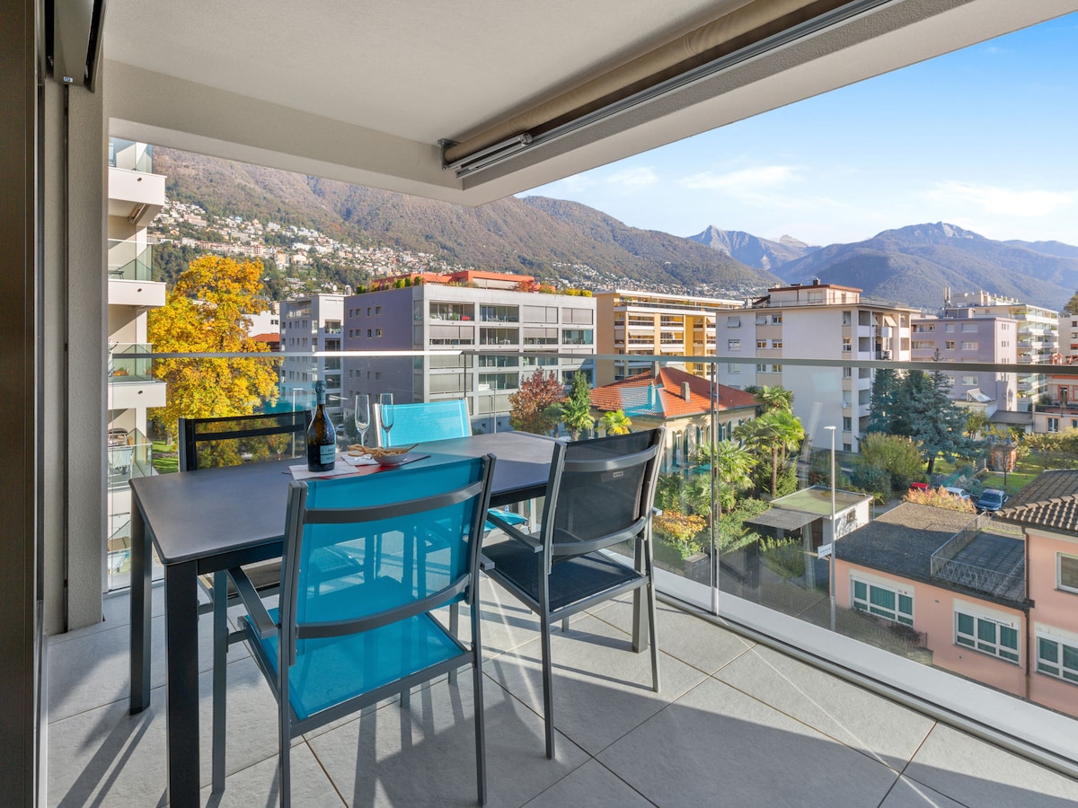 A balcony featuring a dining table surrounded by six chairs is highlighted. The glass railing offers unobstructed views of the surrounding mountains and buildings. Natural light enhances the scene, with greenery visible in the distance, contributing to a serene outdoor space.