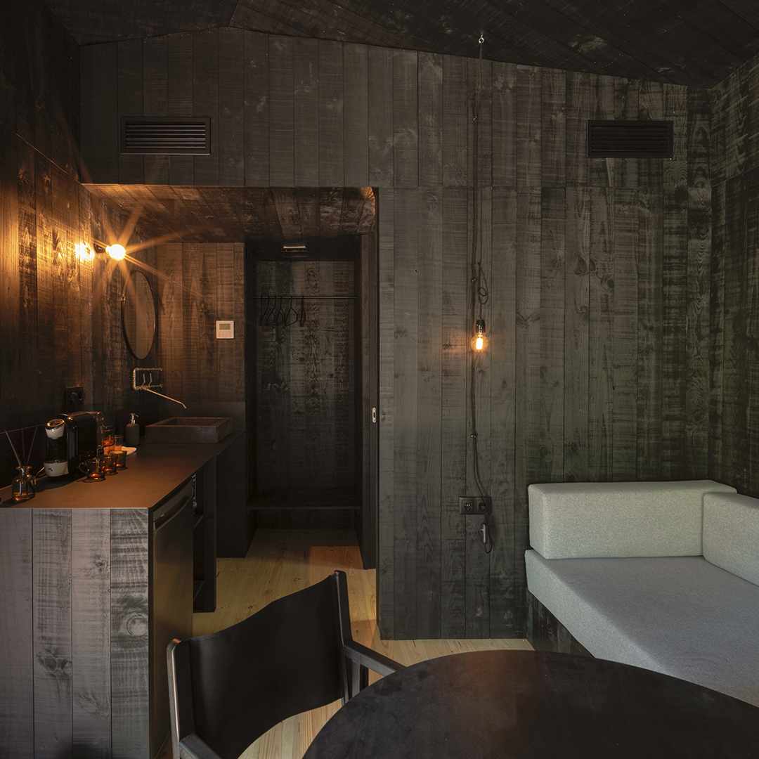 The interior features walls finished in dark wood, creating a cozy ambiance. A small kitchenette is equipped with a coffee machine and a refrigerator. A soft sofa sits adjacent to a round dining table. Warm lighting accents and a doorway leads to the bathroom area.