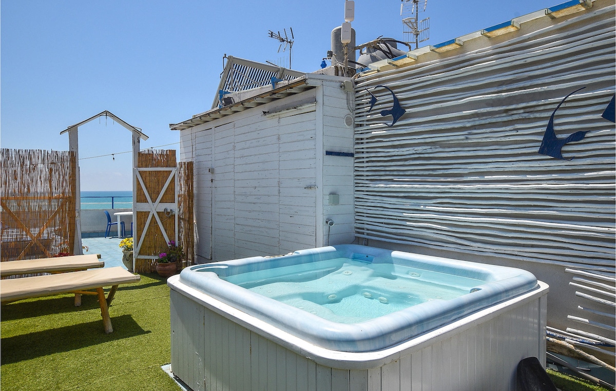A shared outdoor area features a jacuzzi surrounded by lush green grass and sun loungers. The space is enclosed by a white wooden fence, and a clear view of the sea can be seen in the background under a bright blue sky.